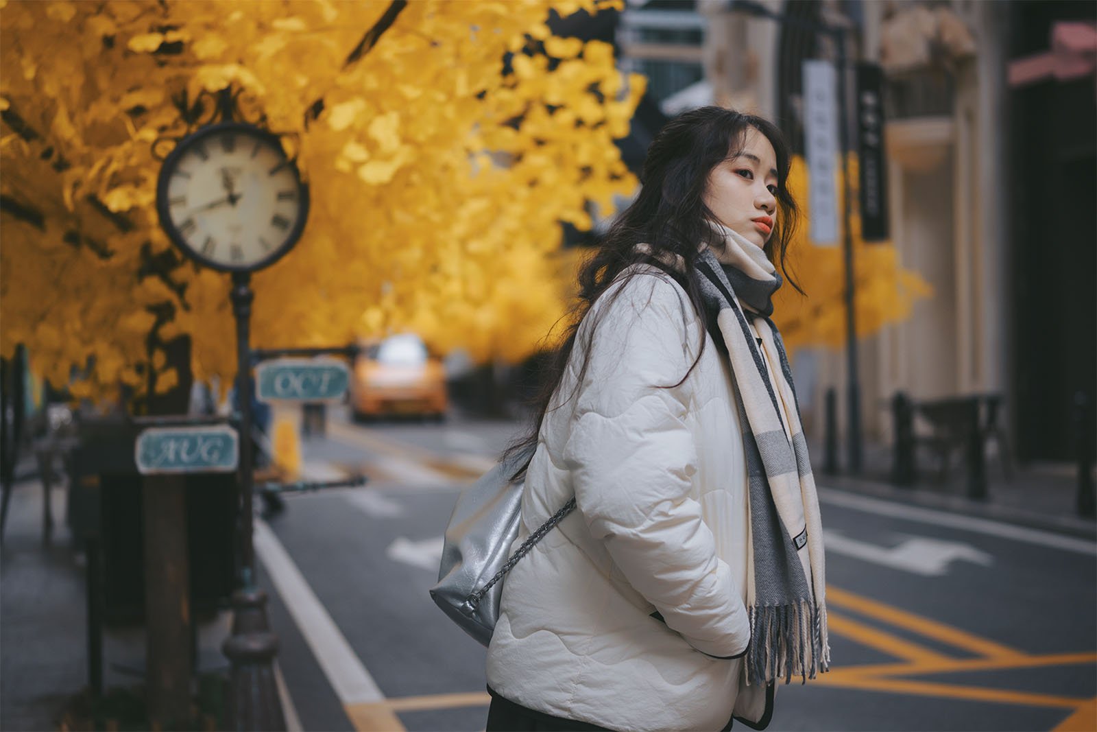 A woman in a white jacket and gray scarf stands by a street, with vibrant yellow autumn trees on one side and a blurry view of a taxi and street clock in the background.