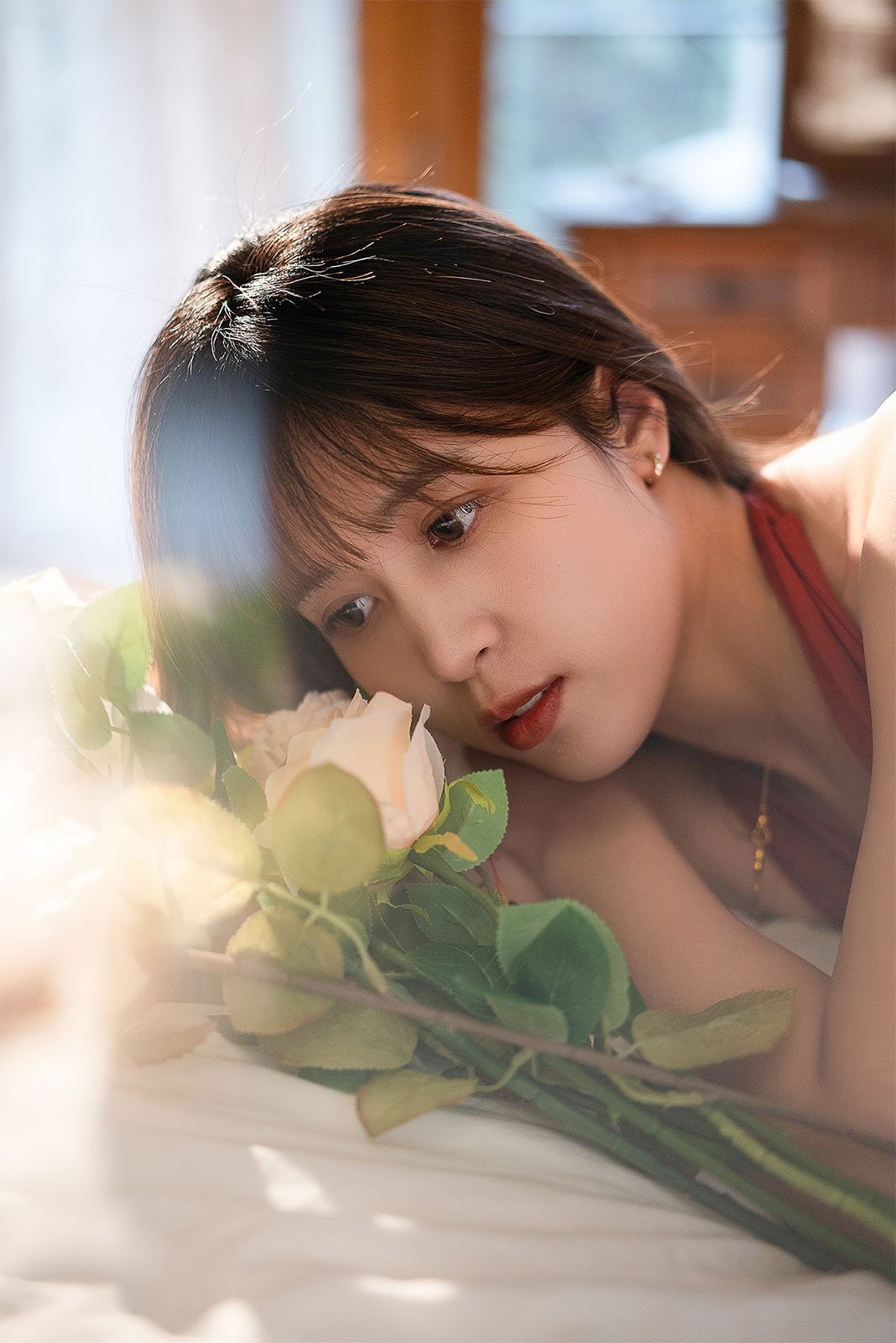 A woman in a red top lies on her side, gazing at a bouquet of white roses with sunlight filtering through a window, creating a warm, serene ambiance.