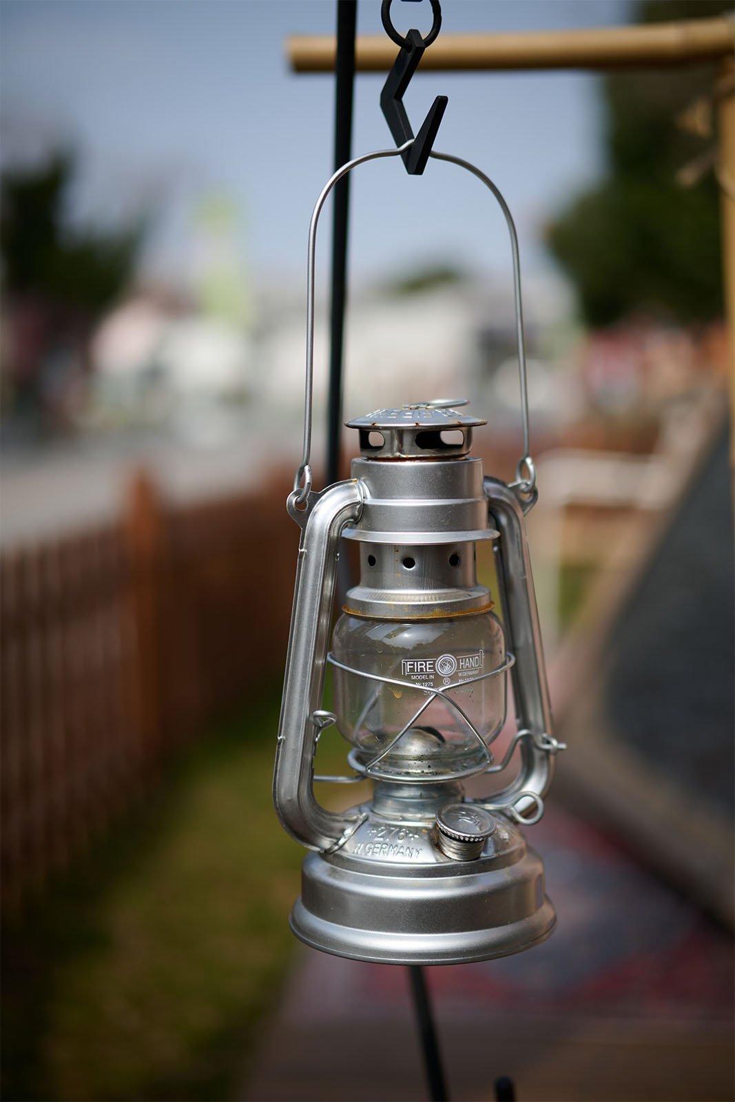 A close-up of a vintage style, silver feuerhand lantern hanging from a metal hook outdoors, with a softly blurred background of a street scene.