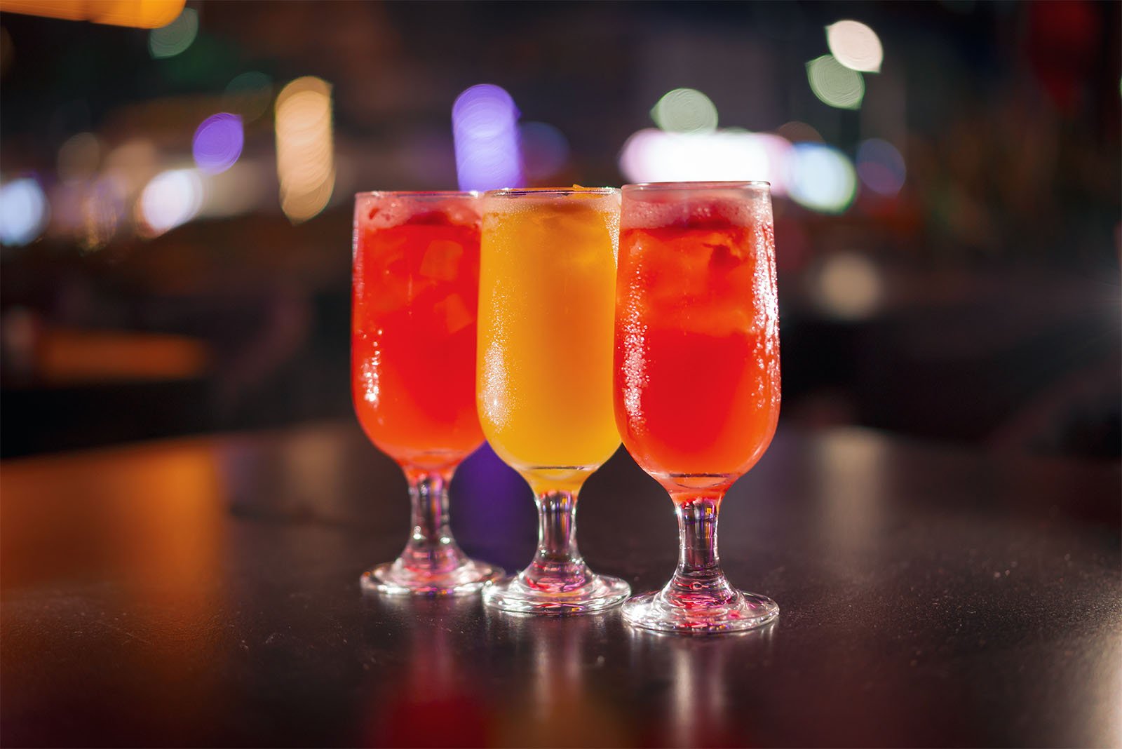 Three colorful cocktails in stemmed glasses on a bar counter, illuminated by soft background lighting with blurred lights.
