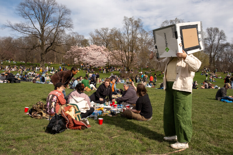 A person wears a homemade pinhole projector next to a group on sitting on the ground.