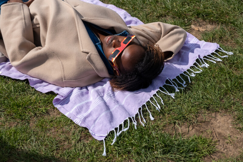 A person lays on a blanket in the grass wearing eclipse glasses.