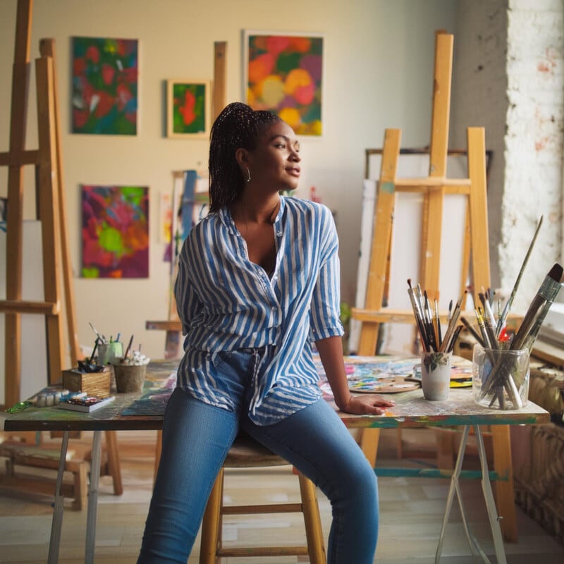 A woman sitting on a stool in an art studio, surrounded by colorful paintings and art supplies, wearing a blue striped shirt and jeans, smiling gently as she looks to the side.