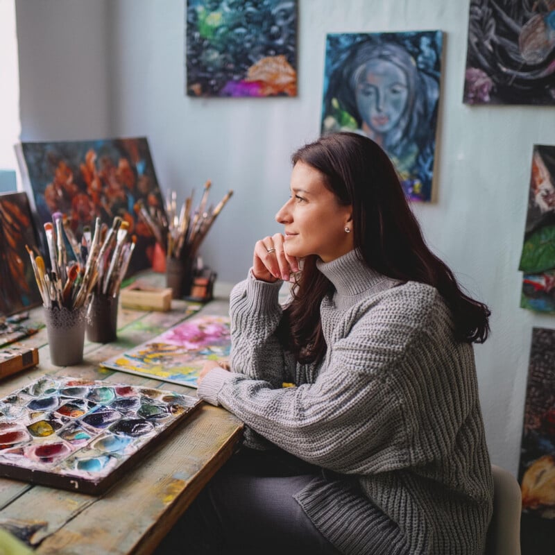 A female artist in a gray sweater sits pensively at a colorful painting workspace filled with brushes and art supplies, surrounded by vibrant paintings.