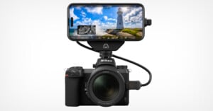 A nikon camera with a large lens connected to a smartphone mounted on top, displaying a lighthouse scene on the phone screen, set against a neutral white background.