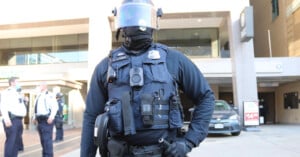 A police officer in full tactical gear, including a helmet with a visor and bulletproof vest, stands in front of a building entrance.