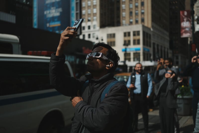 A man wears eclipse glasses, taking a photo with his phone.