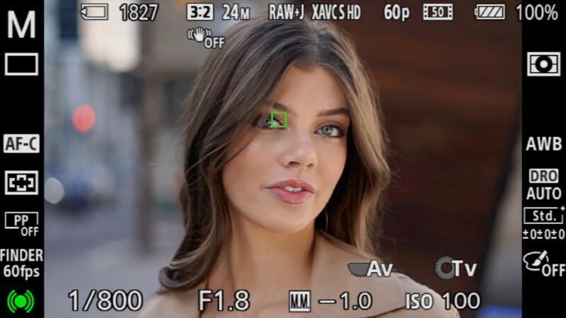 A close-up of a young woman captured on a camera's viewfinder screen, displaying various photography settings such as focus mode, aperture, and iso. she has light makeup and a subtle smile.