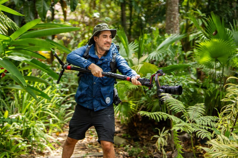 A male photographer in a jungle setting, wearing a hat and carrying a camera with a long lens mounted on a tripod over his shoulder. he looks to his side, smiling as he walks through lush greenery.