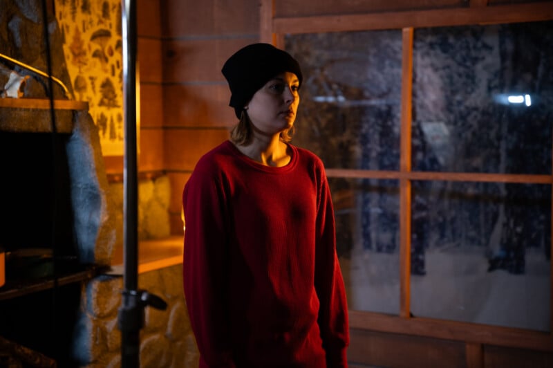 A woman in a red sweater and black beanie stands inside a dimly lit room, looking contemplative. the room has a large window showing a snowy landscape outside.