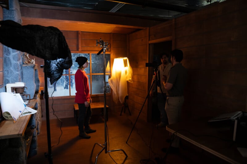 Two people having a conversation on a film set with another person checking the camera setup. the scene is dimly lit and takes place in a room with wooden walls.
