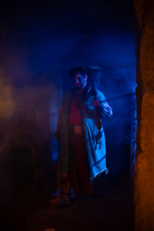 A person wearing traditional attire holds a staff, standing amidst blue fog in a dimly lit, mysterious setting.