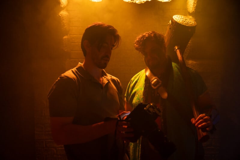 Two men stand in a dimly lit room with an orange glow, one holding a camera and the other holding a sledgehammer, both looking intently to the side.