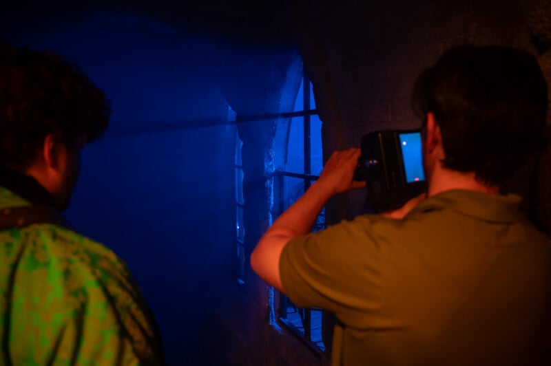 A person is capturing a photo of another individual standing in front of a dimly lit, blue-colored atmospheric scene with a smartphone. the setting appears eerie and mystical.