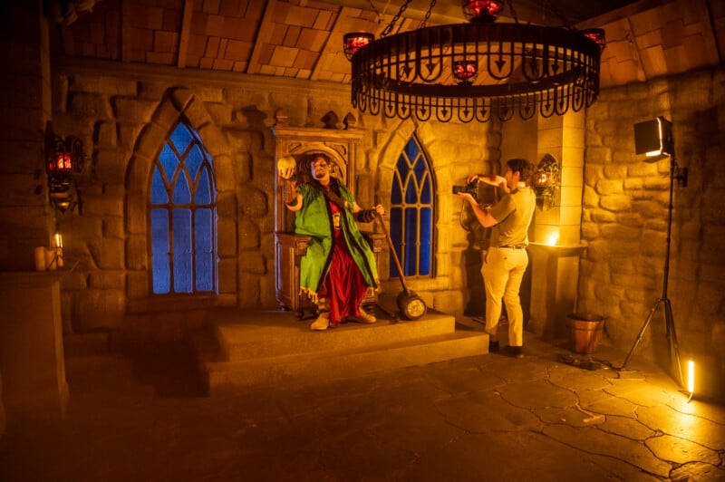 A lively scene inside a castle chamber where an actor in colorful medieval costume performs dramatically on a small stone stage, while another person captures the performance on a digital camera. stained glass windows enhance the ambiance.