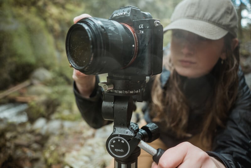 A woman with a cap is adjusting a sony camera mounted on a tripod in a forest setting. the focus is on the camera and the woman appears slightly blurred in the background.