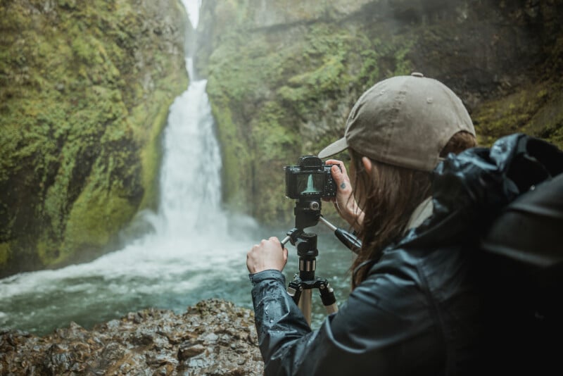 A person wearing a cap is photographing a waterfall using a camera on a tripod, amidst vibrant greenery and mist.