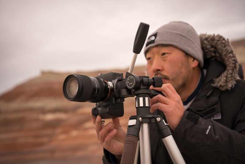 A photographer with an asian descent, wearing a beanie and jacket, focuses intently on adjusting a camera mounted on a tripod, with soft focus desert hills in the background.