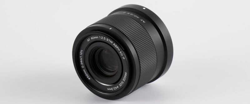 A black camera lens is displayed against a white background, showing focus and aperture details around the front element.