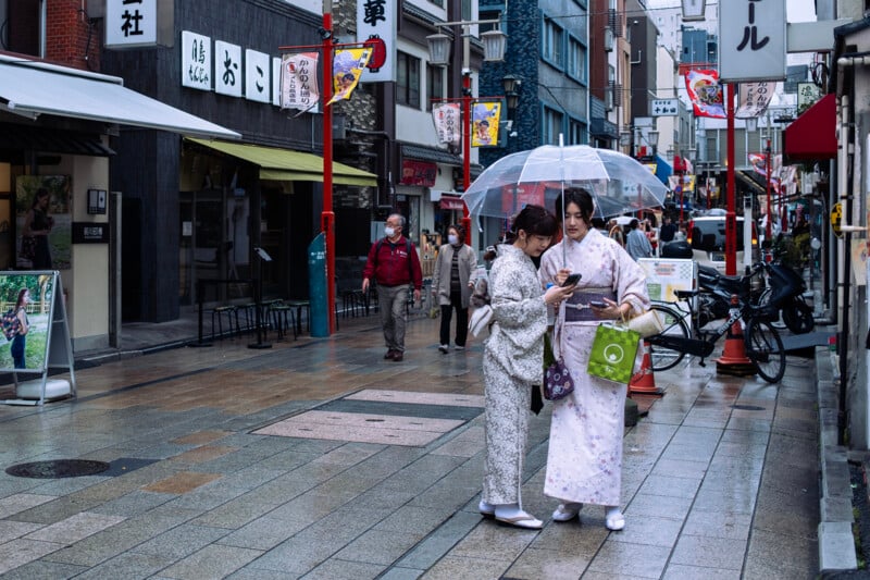 Two women in traditional kimonos check a smartphone under an umbrella on a rainy day in a bustling street with japanese signs and bicycles.