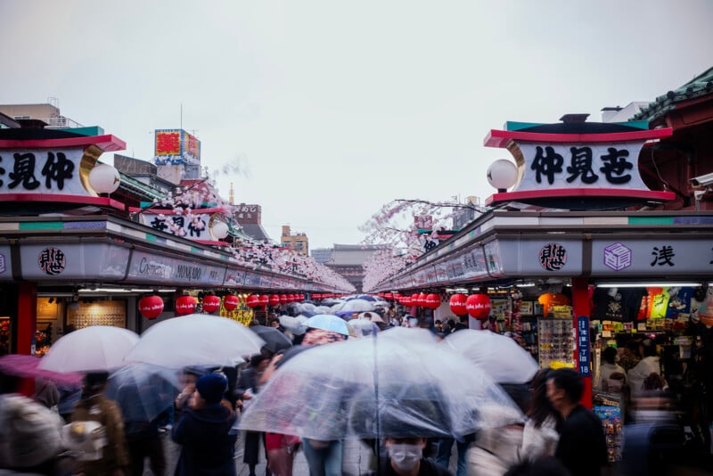 A bustling market street in japan with crowded shoppers carrying umbrellas. red and white signs in japanese above shopfronts. a lively, blurred sense of motion under a cloudy sky.