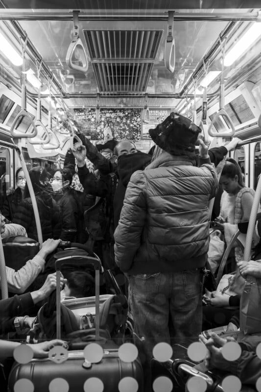 Black and white image of a crowded bus interior with passengers standing and sitting, some holding onto overhead handles, and a prominent figure wearing a puffy jacket and a hat.