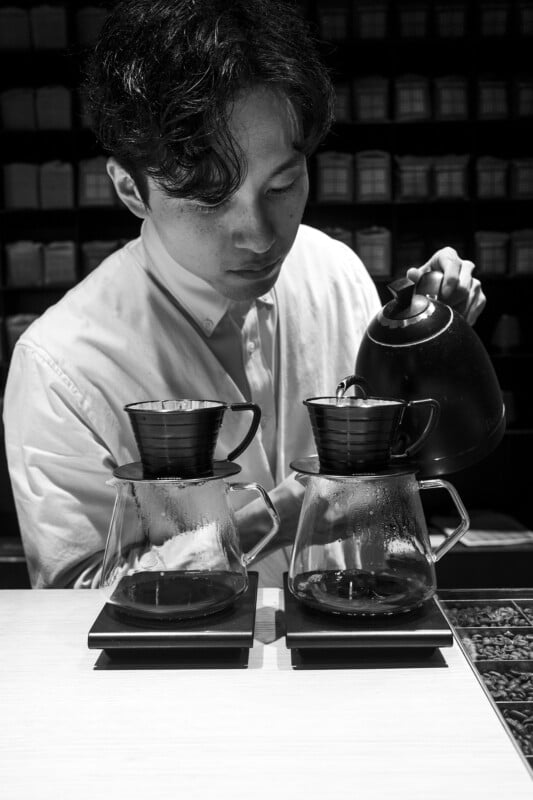 A barista concentrating on pouring hot water from a kettle into two coffee drippers placed atop glass carafes, in a stylish monochrome setting.