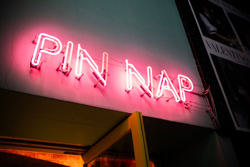 Neon sign saying "pin nap" in pink letters mounted on a wall, casting a warm glow in a dimly lit environment.