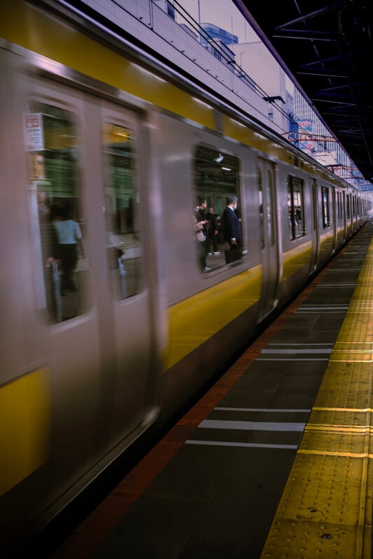 A blurred motion shot of a moving train at a station with passengers visible through the windows. the station platform has a yellow safety line and is partially covered.