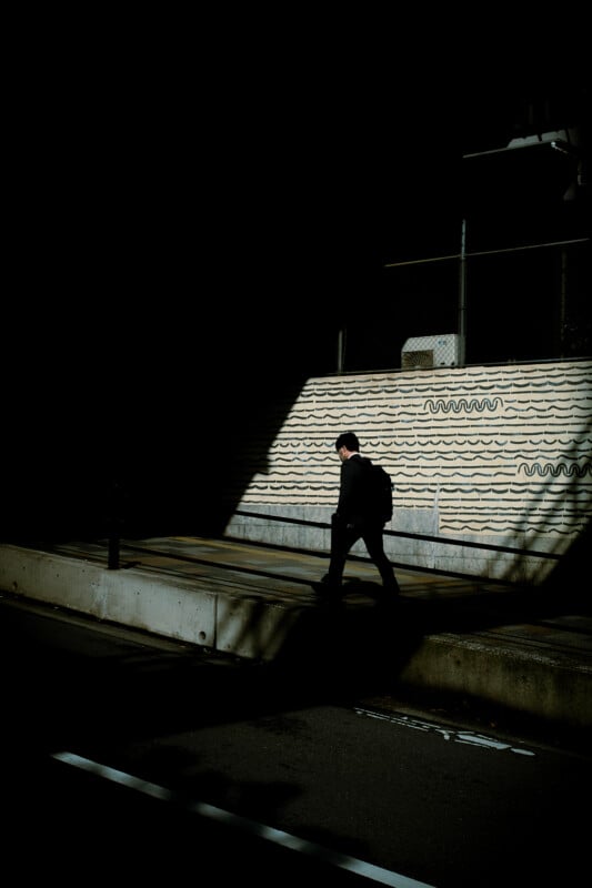 A man walks swiftly past a wavy-patterned wall in a shadowy street, with sunlight highlighting his path and the upper portions of the wall.