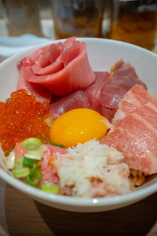 A bowl of chirashizushi featuring slices of fresh sashimi, including tuna and salmon, a mound of salmon roe, a raw egg yolk, and shredded crab meat, garnished with green onions.