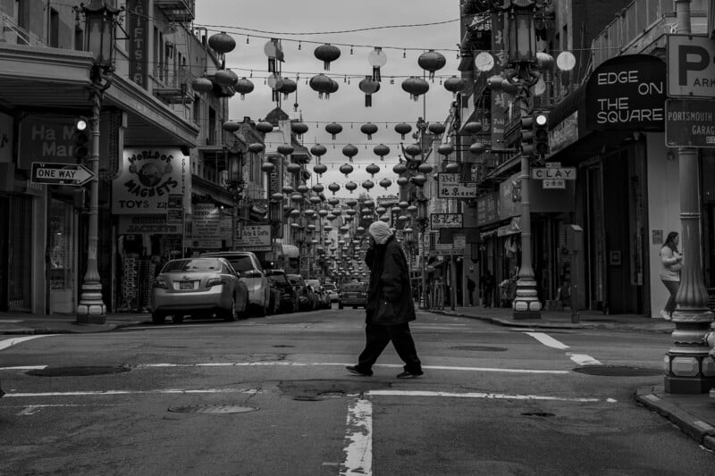 A black and white photo of an elderly person crossing a street lined with hanging lanterns and various shop signs in an urban setting, capturing a tranquil moment amidst city life.