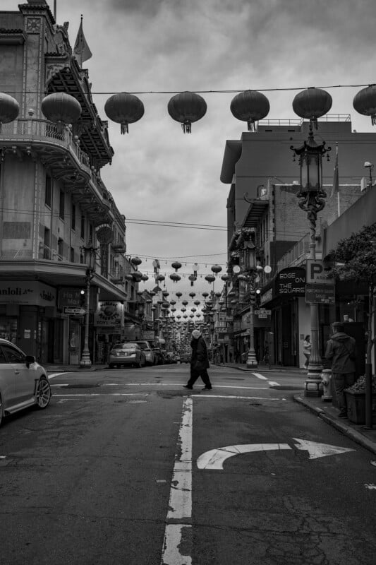 A black and white photo of a person crossing a street in an urban area decorated with hanging lanterns, with classic architecture on either side.