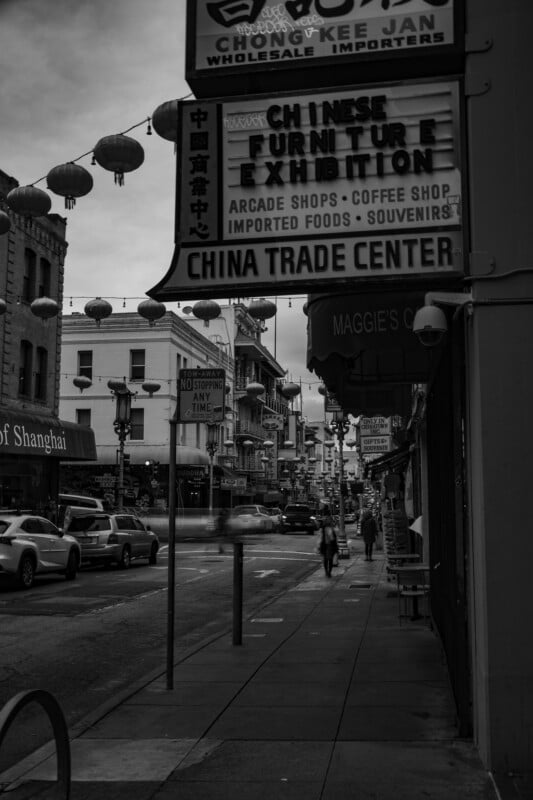 A monochrome urban street scene in chinatown with various signs displaying chinese characters, including "chinese furniture" and "china trade center." pedestrians and shopfronts line the busy street.