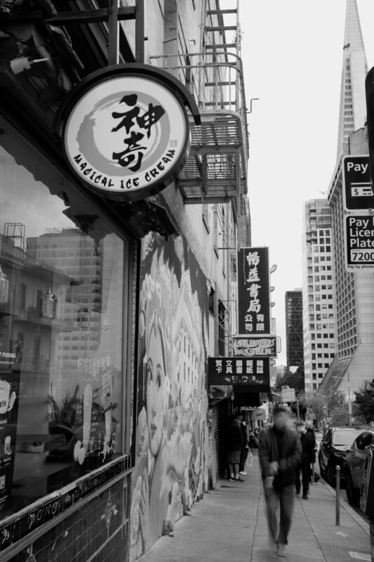 Black and white photo of a city street with a "magical ice cream" shop sign in the foreground. a mural decorates the building, and a blurred figure walks by. skyscrapers are visible in the background.