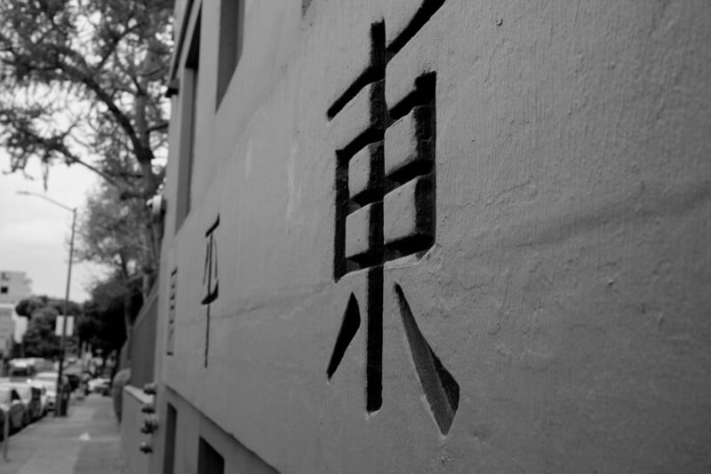 Black and white photo of a large chinese character painted on a textured city wall, with blurred street scenery in the background.