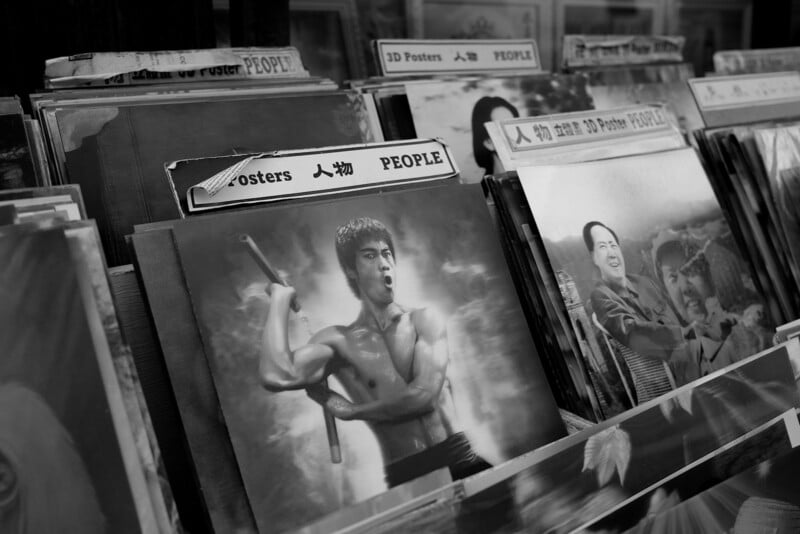 Black and white photo of a rack displaying various posters including a prominent one of a man with a martial arts stance. other posters feature people and celebrities.