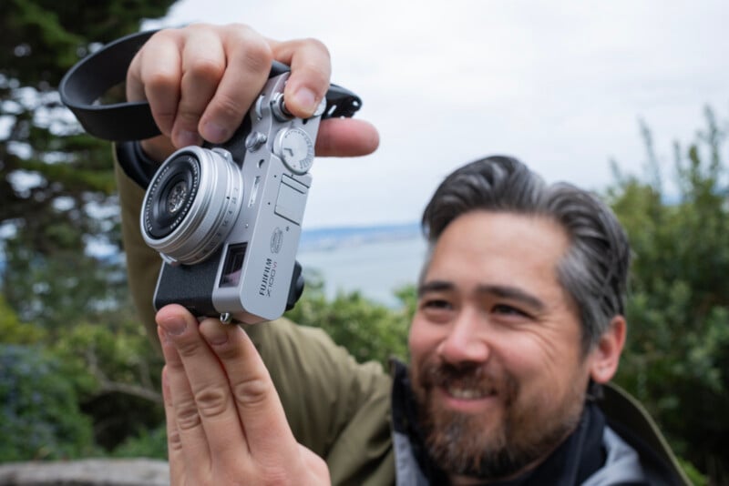A smiling asian man holding a silver digital camera, focusing it towards the viewer, with trees and a blurred coastal background.