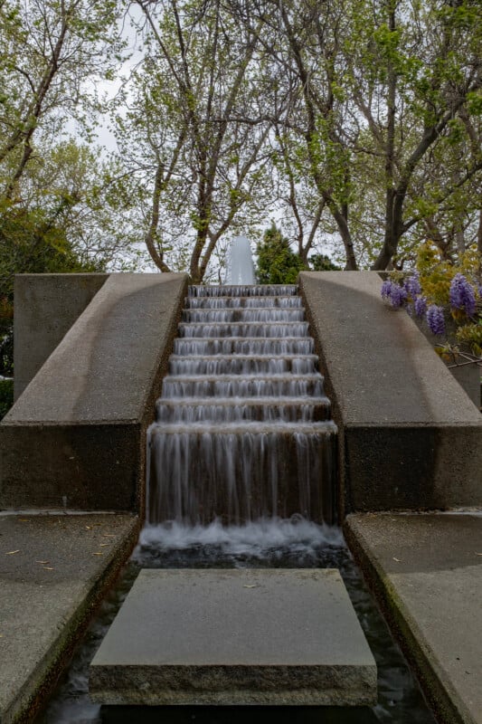 Concrete stepped waterfall in a park with flowing water, surrounded by lush green trees and purple flowers, under an overcast sky.