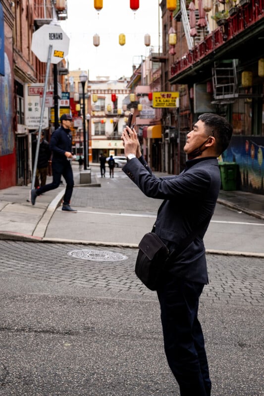 A man in a dark suit takes a photo with his smartphone in a vibrant alley decorated with red lanterns, while another man walks away in the background.
