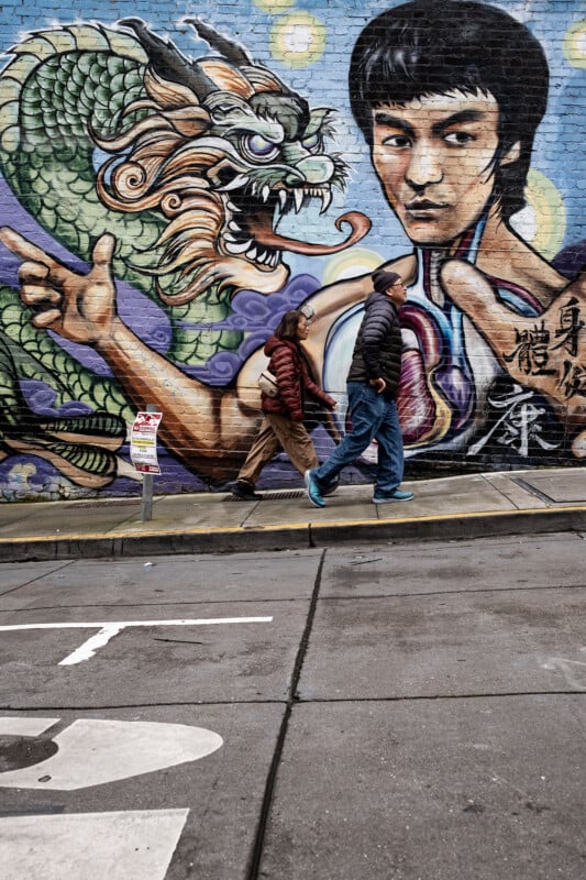 Two people walk by a vibrant street mural featuring a large, detailed image of bruce lee in a fighting stance and a green dragon, set against an urban backdrop.