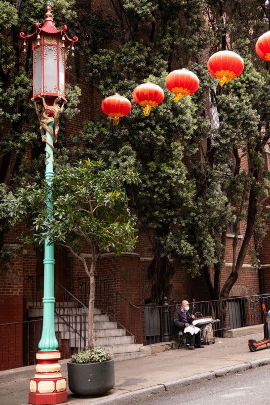 Red lanterns hang above a street in a festive display. an ornate lamp post stands on the left, and an elderly person reads a newspaper on a bench under a canopy of trees.