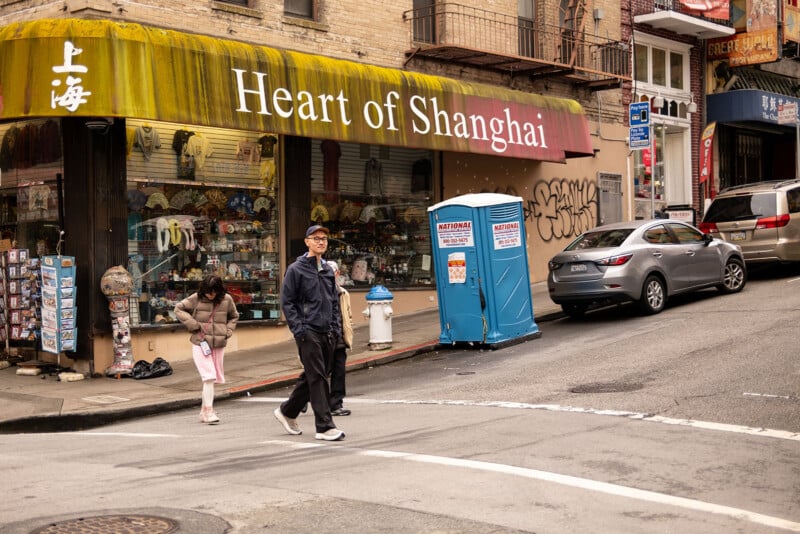 A man in a black jacket walks past the "heart of shanghai" store on a bustling city street with parked cars, various storefronts, and pedestrians in the background.