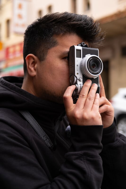 A man using a silver vintage camera to take a photo in an urban setting. he is wearing a black hoodie, focusing intently through the camera's viewfinder.