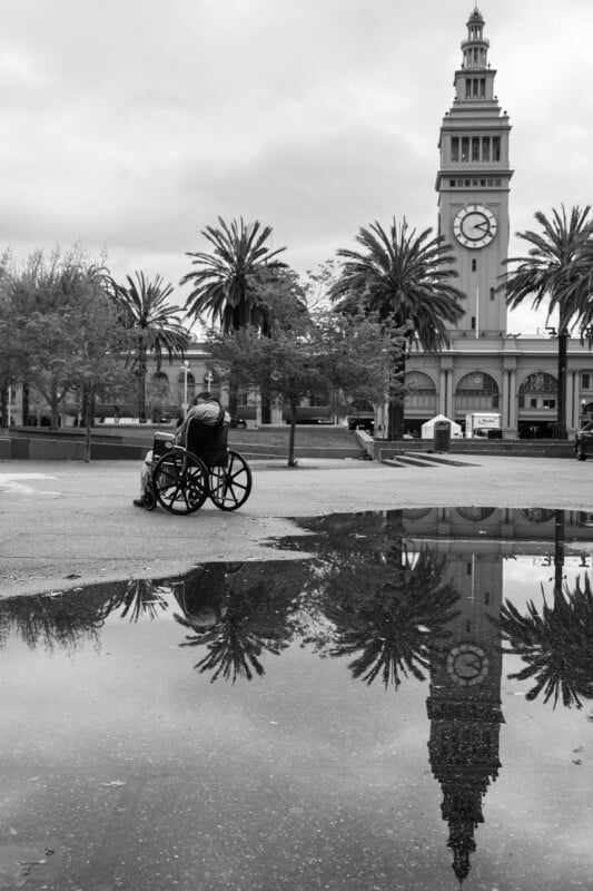 A person in a wheelchair is reflected in a puddle, looking toward a clock tower surrounded by palm trees in a cloudy, open plaza.