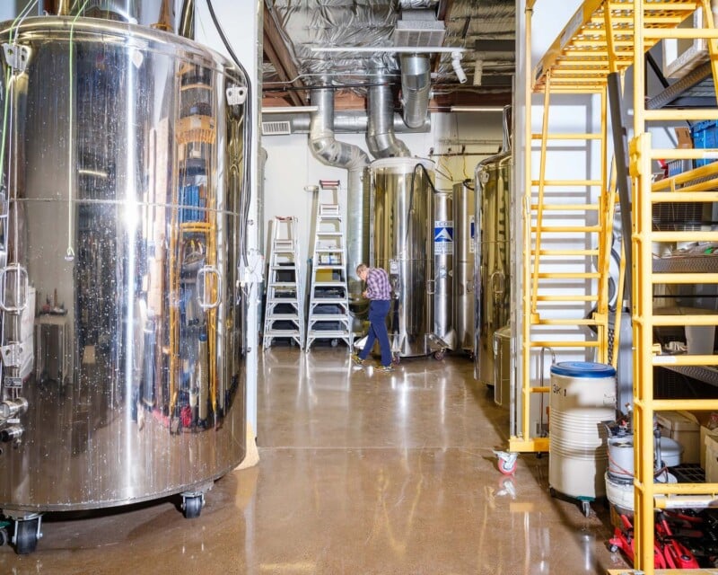 A brewery interior with large stainless steel fermentation tanks, a person cleaning the floor, and shelves stocked with supplies.