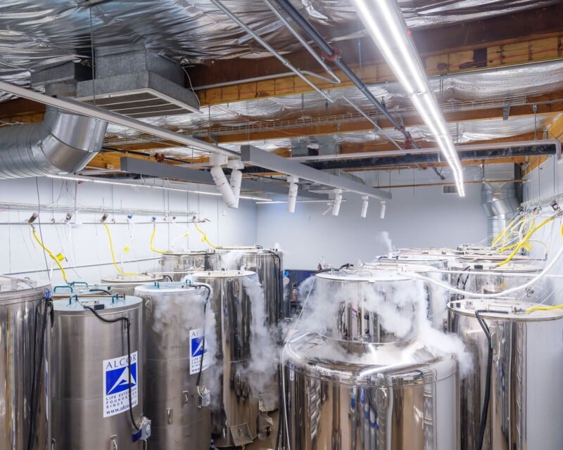 A modern brewery's fermentation room with several large stainless steel tanks emitting vapor, connected by numerous overhead pipes and tubes, under a ceiling lined with exposed insulation.