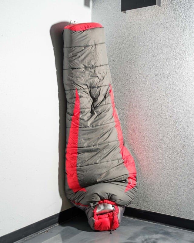 A mummy-style sleeping bag in grey and red, propped up vertically against a white wall, positioned in a corner with contrasting light and shadow.