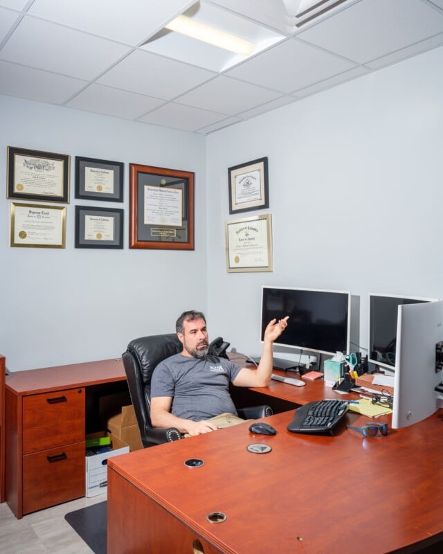A man in a gray t-shirt sits reclined in an office chair, gesturing while talking. his desk has dual monitors and is surrounded by framed certificates on the walls.