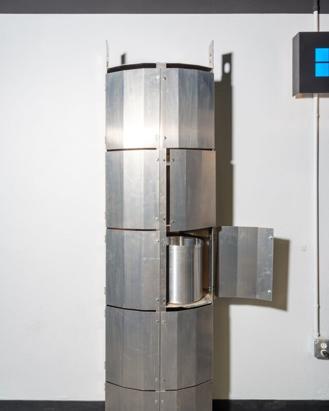 A tall, metallic, segmented sculpture resembling a vertical column with various hinged panels opened at different angles, displayed in a gallery setting with a white wall background.